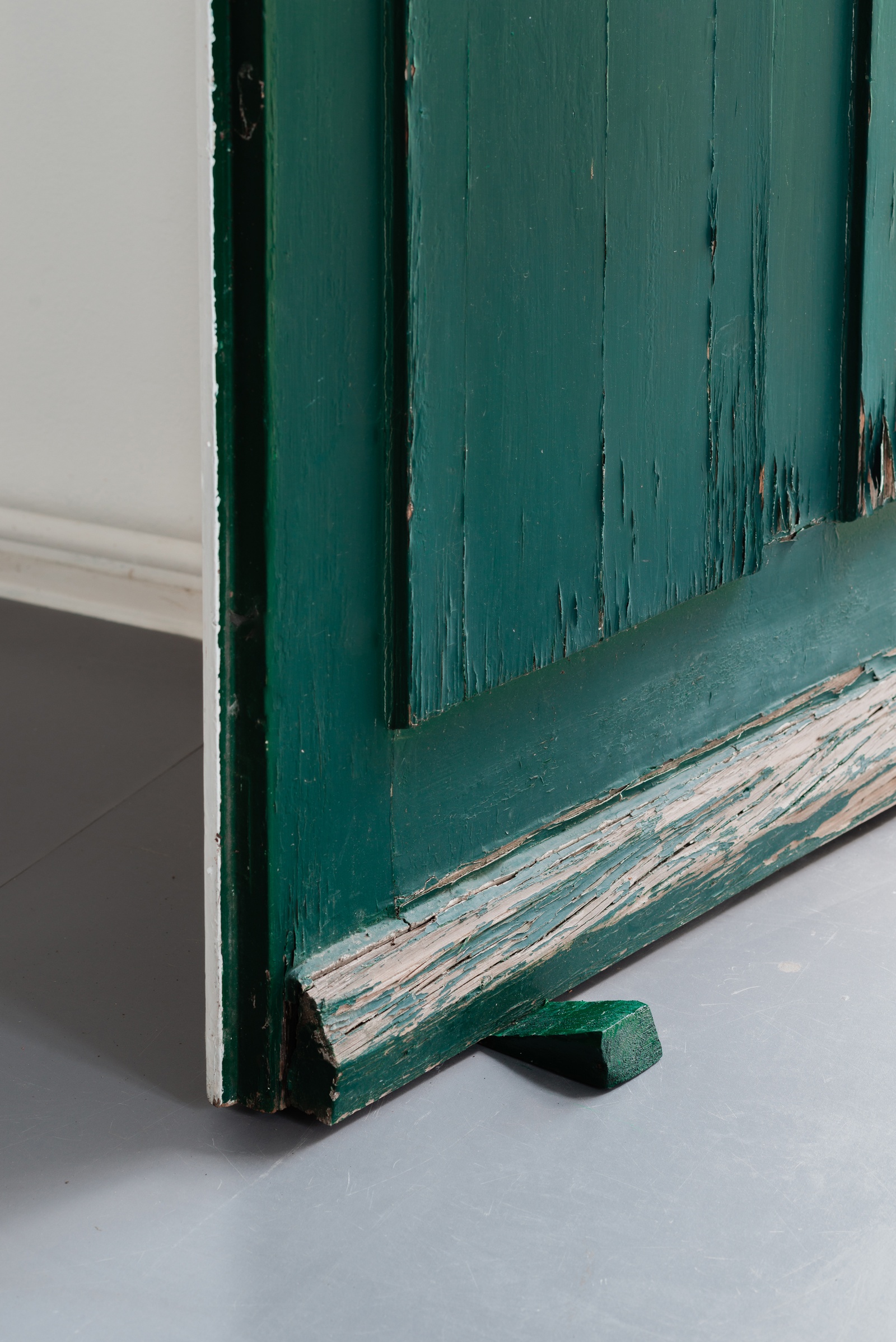 [A detail of the left corner of green wooden door where the doorstop rests. The wooden doorstop is the same green color as the door. The door shows layers of peeling paint, and the cracked wood’s rough texture contrasts with the smooth, gray floor beneath. The light falls in from the left, casting subtle shadows that accentuate the worn edges.]