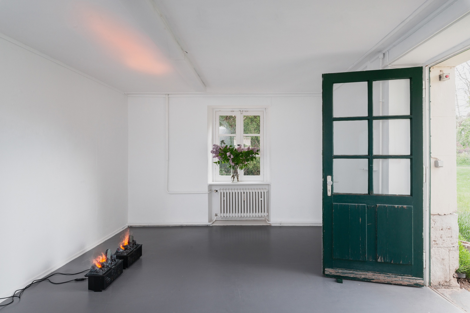 [In a white exhibition space, a green door stands wide open on the right, its swing obstructed by a wooden doorstop of the same color. On the back wall, centered beneath a heater and in front of the window, there is a flower arrangement on a ledge. The arrangement features tree branches with purple blossoms and dark green leaves. On the left wall, opposite the flowers, are two identical objects resembling fireplaces, mounted on electrical cabinets. The wood is charred, with the flames glowing brightly.]