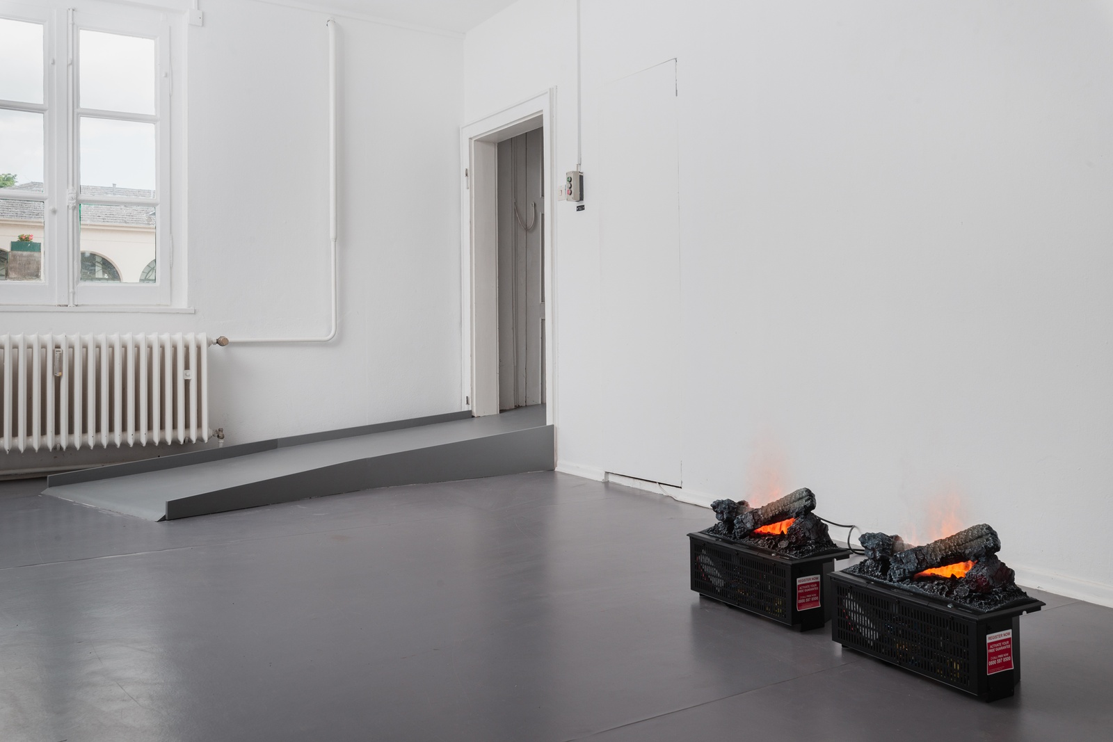 [In a white space with gray painted floors, a raised doorway in the corner is made accessible with a gray ramp. On the right side of the ramp, parrallel to the wall are two identical small black boxes sit directly on the floor. On their top, identical configurations of logs appear burning in flames. Through small slits in the boxes, wires and electrical components are visible. On the right side of each boxes, there&#x27;s a sticker that reads, “REGISTER NOW; ACTIVATE YOUR FREE GUARANTEE; CALL FREE NOW; 0800 597 8500.”]