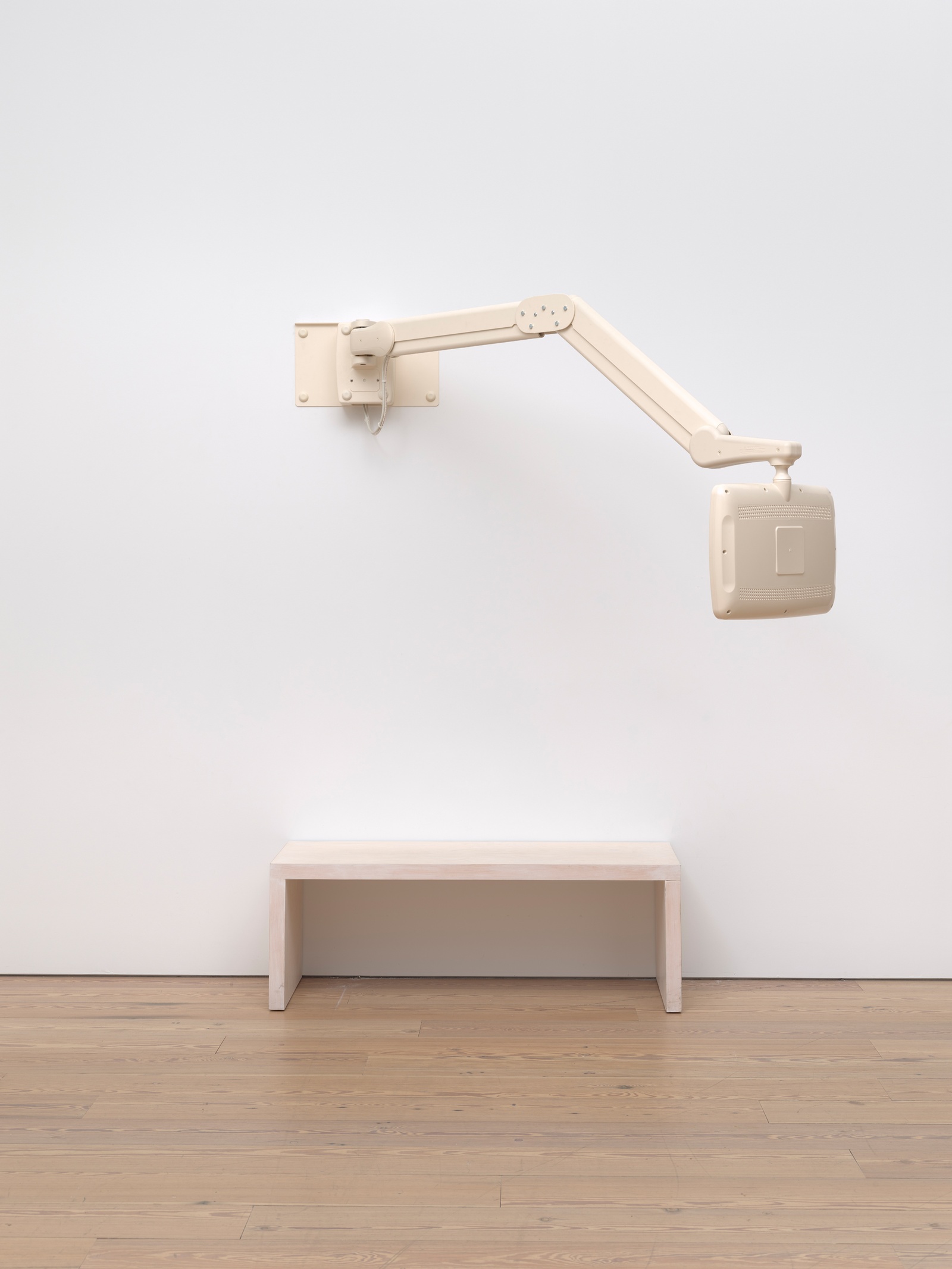 [In a white space with a light wood floor, a beige hospital television monitor attached to a beige mechanical arm is mounted to the wall and extends outward. Below it, a matching wooden bench sits against the wall. From this perspective, the monitor is facing the wall and the bench.]