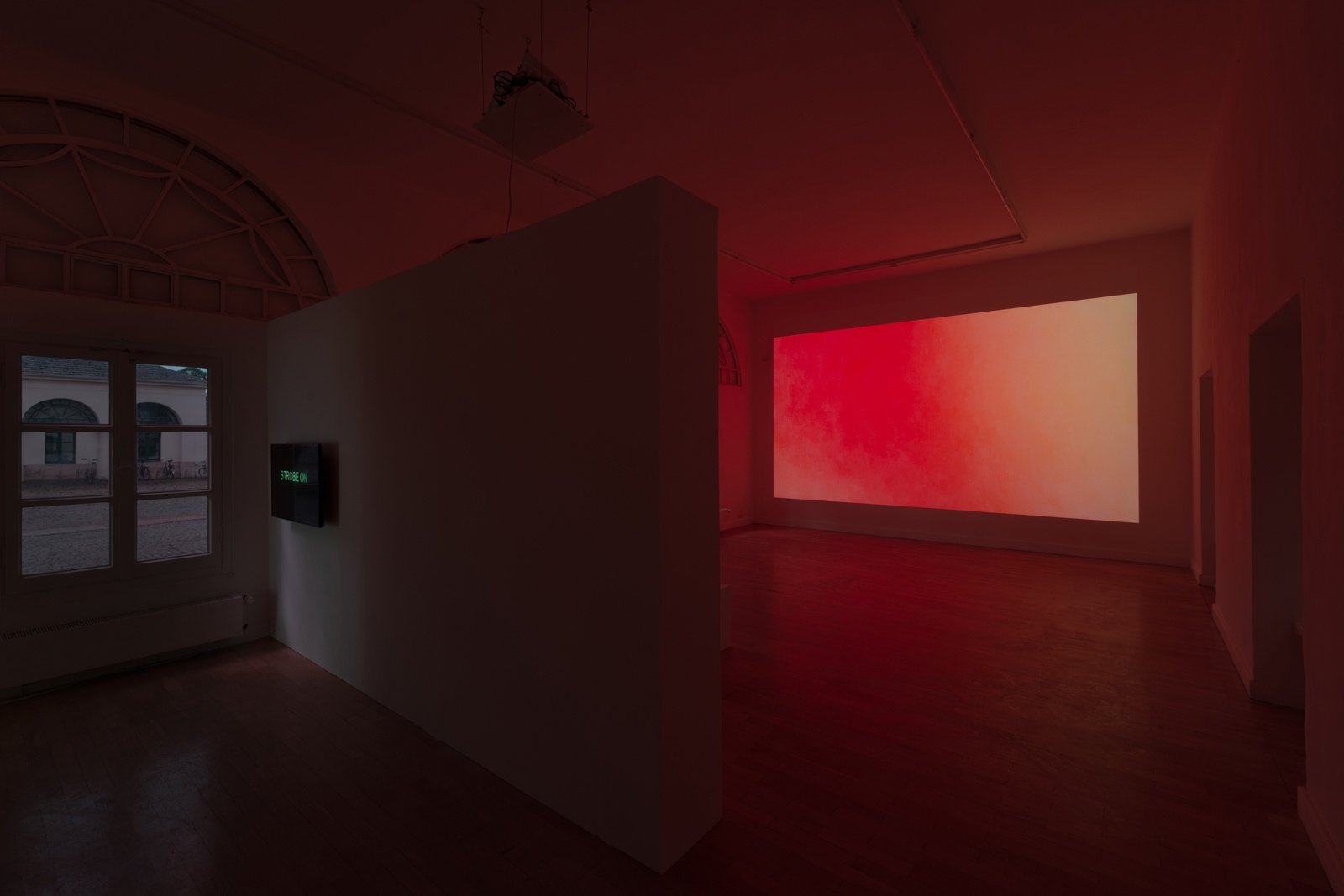 [A dimly lit exhibition space with a warm red glow with wooden flooring and white walls, some of which are used as partitions. A large rectangular projection on one of the walls displays a gradient of red and pink hues, casting a soft light across the space. On the left side, a smaller screen with green text on a black background is mounted on a partition wall near a window with arched frames. The window reveals an outdoor scene with a white building in the background.]