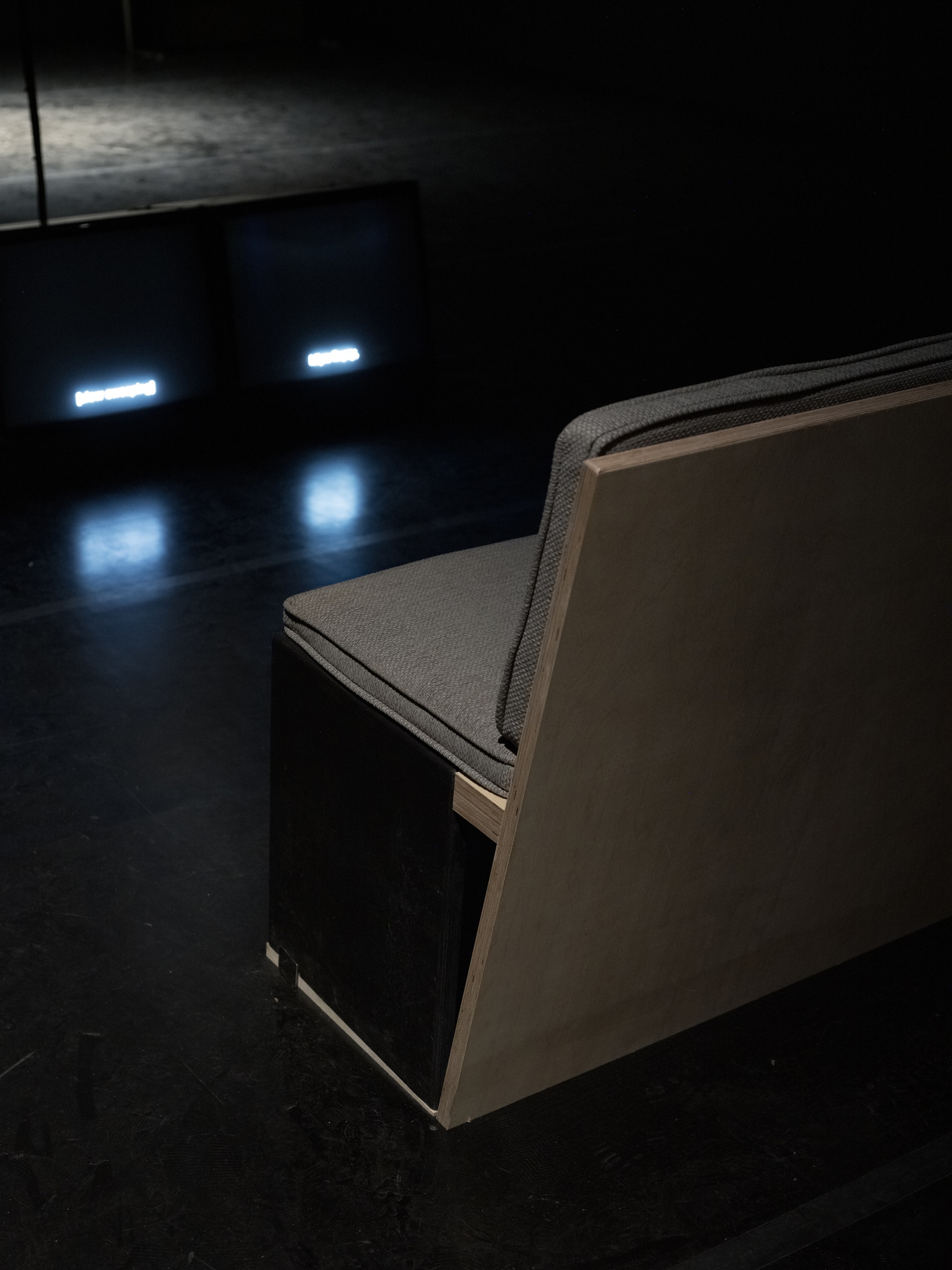 [In a dark room, a close-up view of an exhibition bench is shown, with wooden bases and gray cushioned tops, seen from the top left. On the right side, two side-by-side television monitors on the floor display white subtitles.]