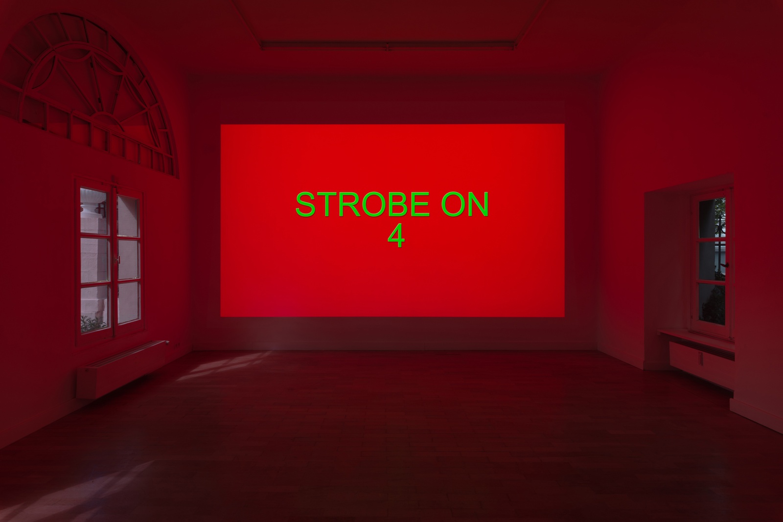 [A dimly lit exhibition space with a large rectangular red screen projection on the wall with a green text, reads: STROBE ON/4. On both side of the screen are windows that reveal outdoor light.]