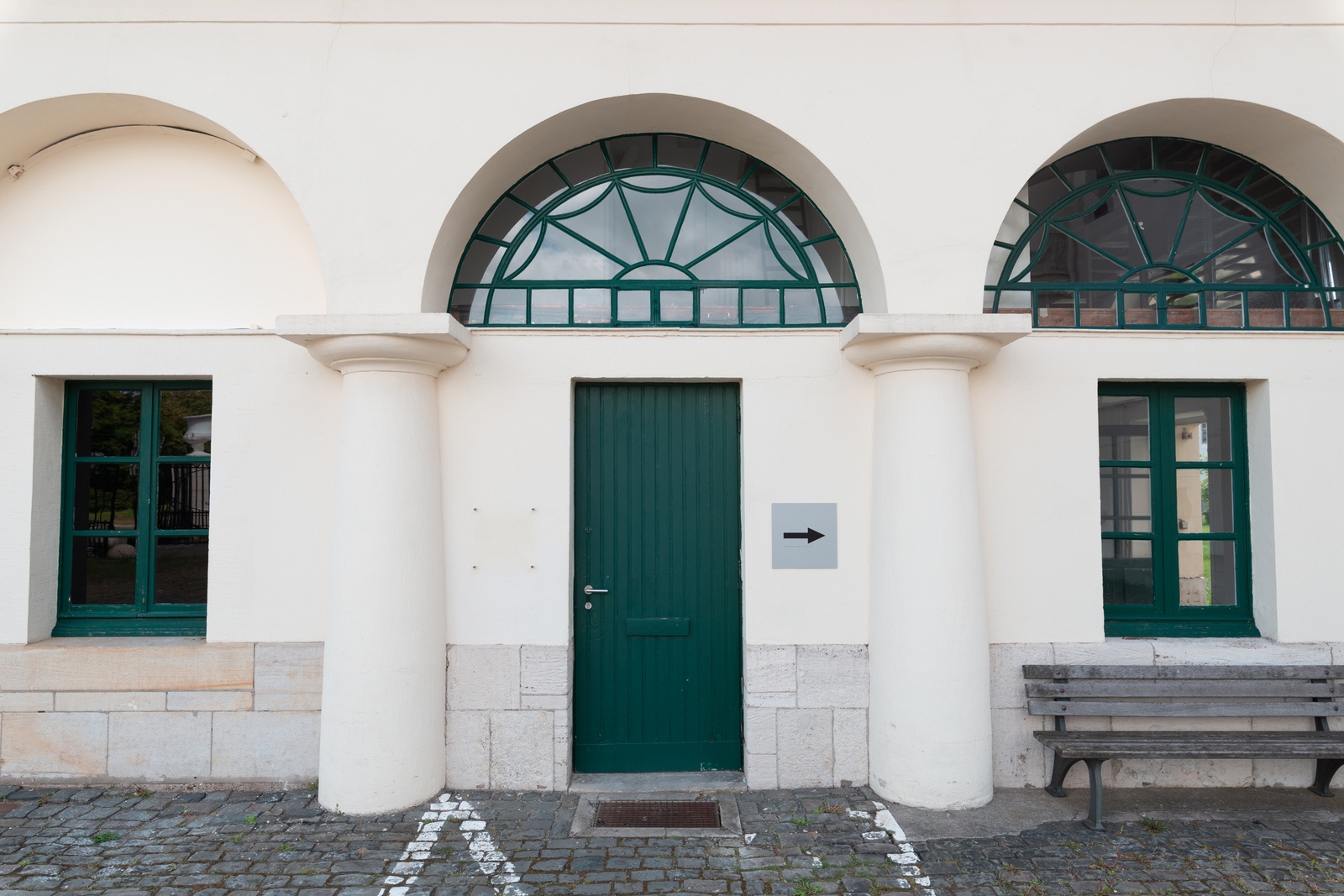 [A building with a symmetrical design features three distinct sections, each framed by large, white columns that support rounded arches. The central section contains a green door with vertical panels, flanked by two white columns. To the right of the door, there is a gray square sign with a black arrow pointing to the right. On either side of the door are windows with green frames. A wooden bench is positioned against the wall beneath the right window. The ground in front of the building is paved with cobblestones, and faint white markings are visible on the pavement near the columns.]