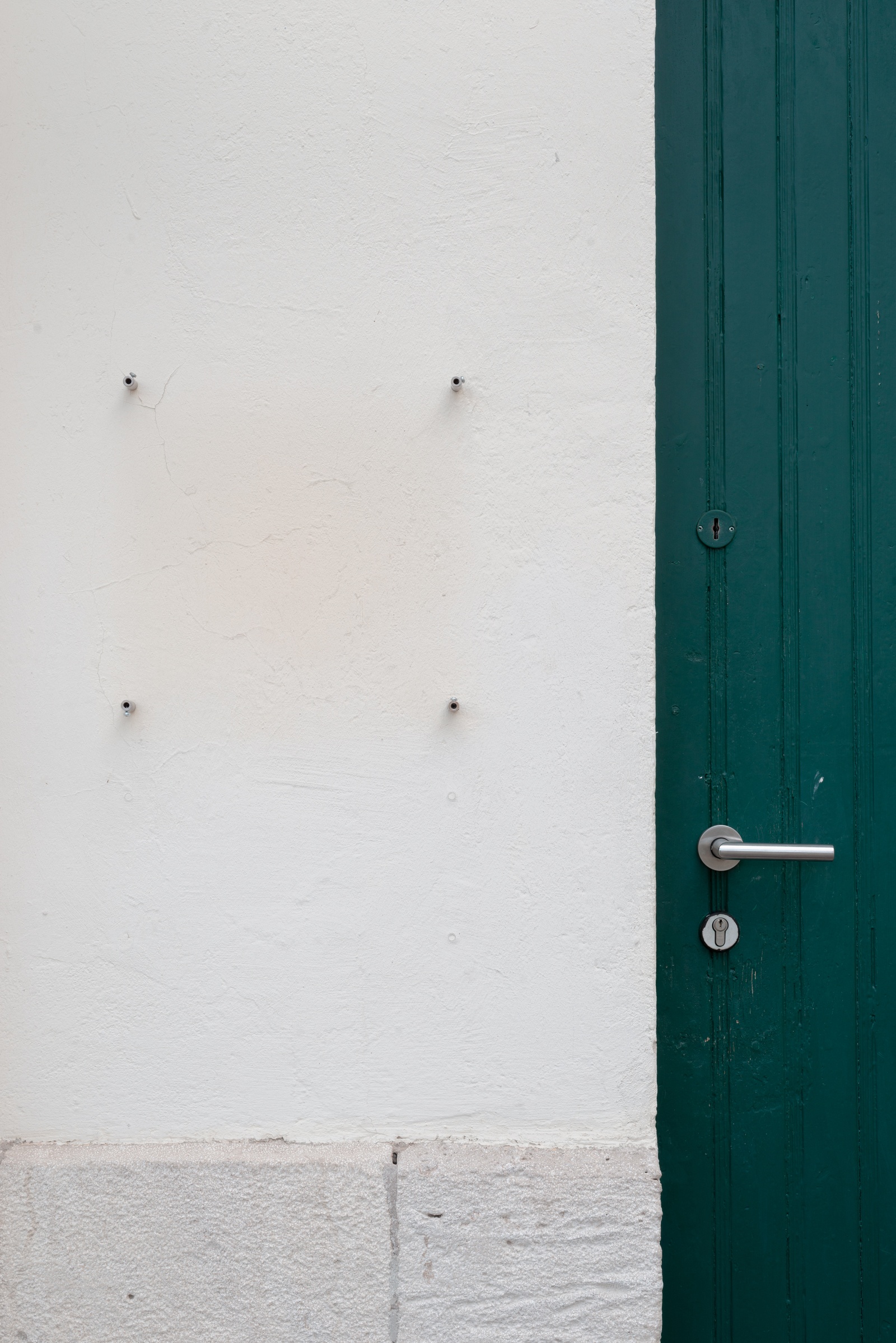 [The image shows a section of a plain, white-painted wall with a green door on the right side. The door is vertical, made of wood, and painted a deep green color. It has a modern silver handle and a round keyhole below it. On the white wall to the left of the door, there are four small metal pegs or screws arranged in a rectangular pattern, two on top and two at the bottom, suggesting that something was once mounted there, like a sign or a plaque. The paint around this area is slightly discoloured, indicating the outline of whatever used to be attached. The bottom part of the wall features a strip of light grey stone.]