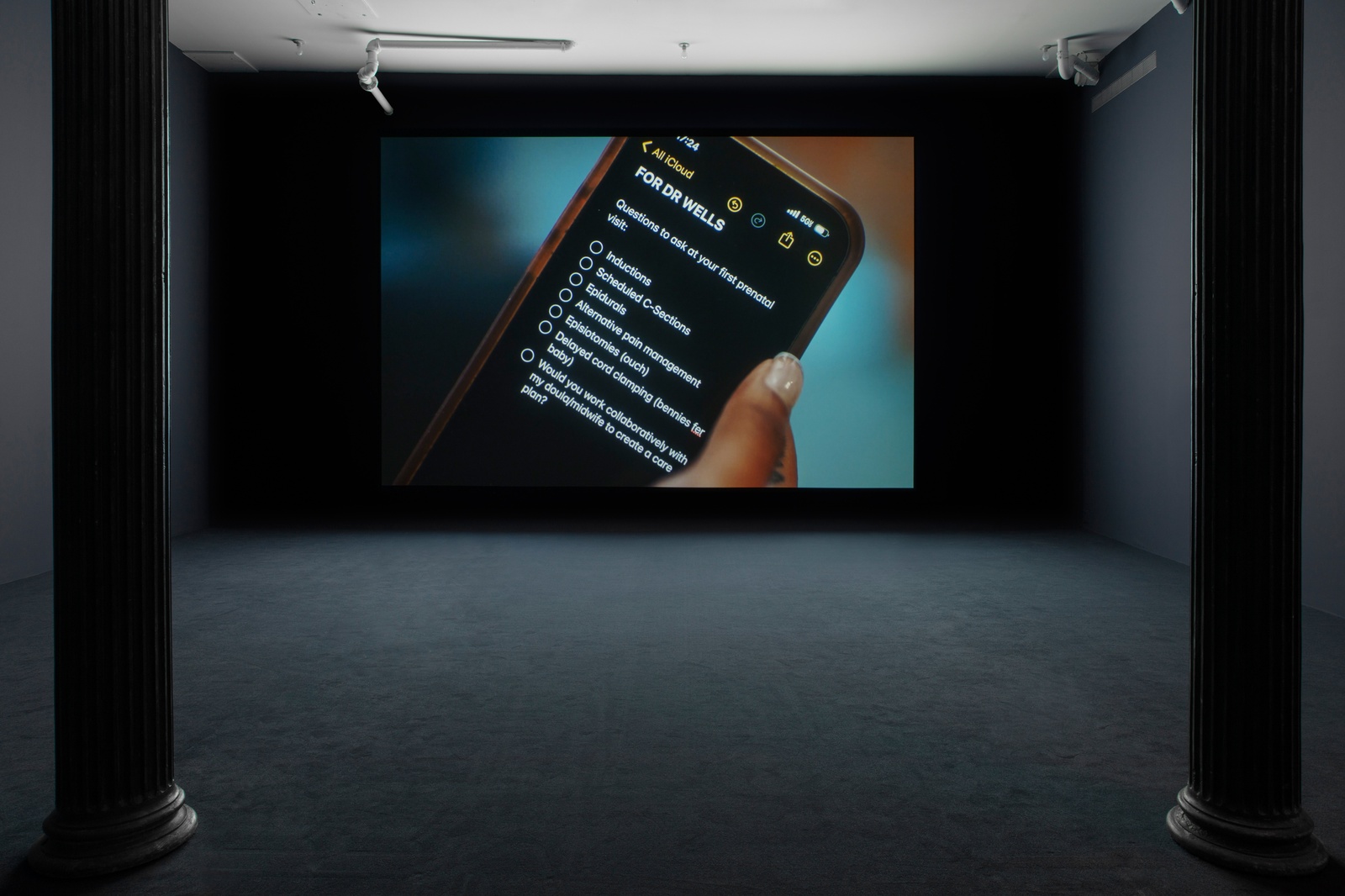 Carolyn Lazard: Two-way. Installation view (Vital, 2025), Artists Space, 2025. Photo: Greg Carideo. Image Courtesy Artists Space, New York. [A darkened gallery space with dark blue walls and dark blue carpeting. A film is projected onto the farthest wall.The projection depicts a brown hand holding an iPhone screen opened to a bullet point list that reads: “FOR DR WELLS / Questions to ask at your first prenatal visit / Induction / Scheduled C-Sections / Epidurals / Alternative pain management / Episiotomies (ouch) / Delayed cord clamping (bennies fer baby) / Would you work collaboratively with my doula/midwife to create a care plan?”]
