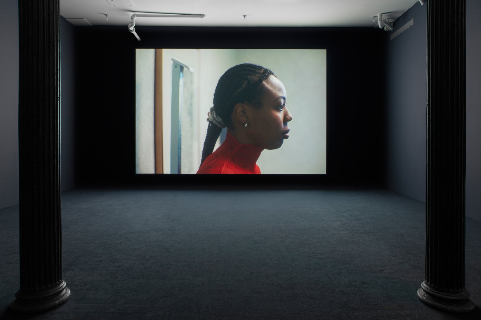 Carolyn Lazard: Two-way. Installation view (Vital, 2025), Artists Space, 2025. Photo: Greg Carideo. Image Courtesy Artists Space, New York. [A darkened gallery space with dark blue walls and dark blue carpeting. A film is projected onto the farthest wall.The projection depicts a black person with long braids in side profile standing in front of a ruler.]