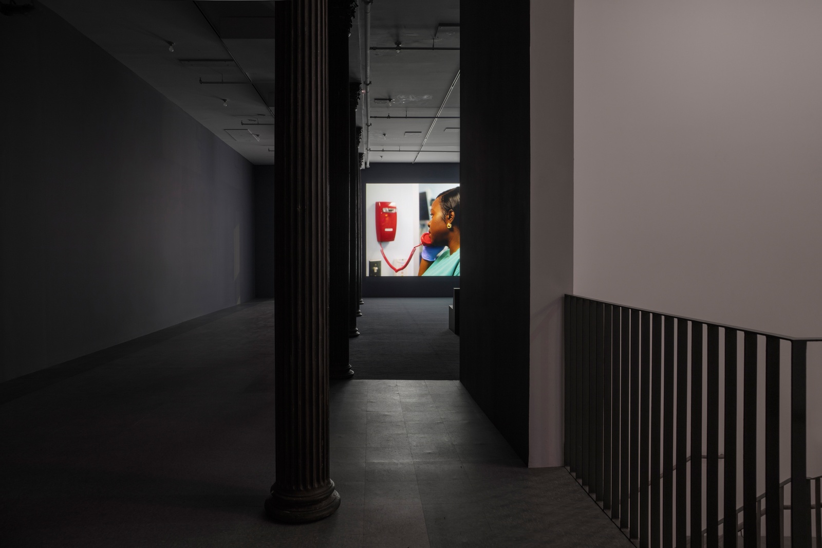 Carolyn Lazard: Two-way. Installation view (Fiction Contract, 2025; Borne, 2025), Artists Space, 2025. Photo: Greg Carideo. Image Courtesy Artists Space, New York. [A darkened hallway leads into an open gallery space with dark blue walls and a combination of light blue linoleum tile and dark blue carpeting. In the background a video is projected onto the farthermost wall. The projection depicts a nurse in green scrubs using a red telephone affixed to the wall.]