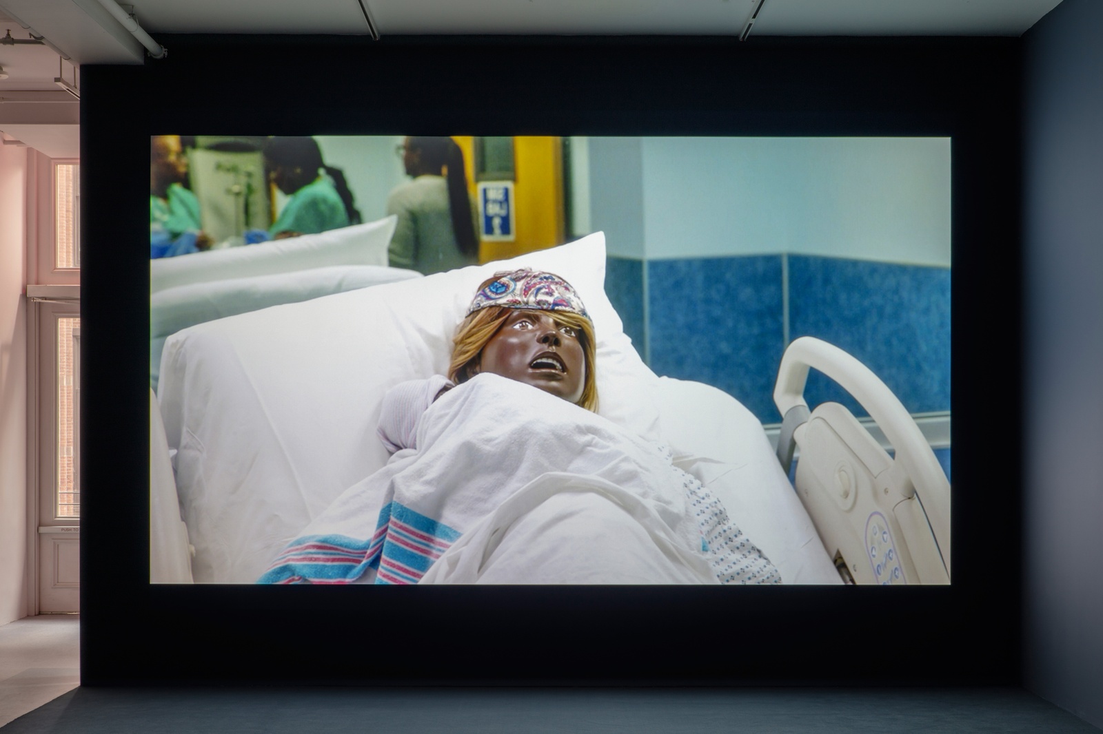 [A darkened gallery space with a video projection centered on a dark blue wall. The projection depicts a close-up of a black birthing mannequin reclined on a hospital bed in a blue patient room. A newborn mannequin swaddled in a hospital receiving blanket lays on the birthing mannequin’s chest.]