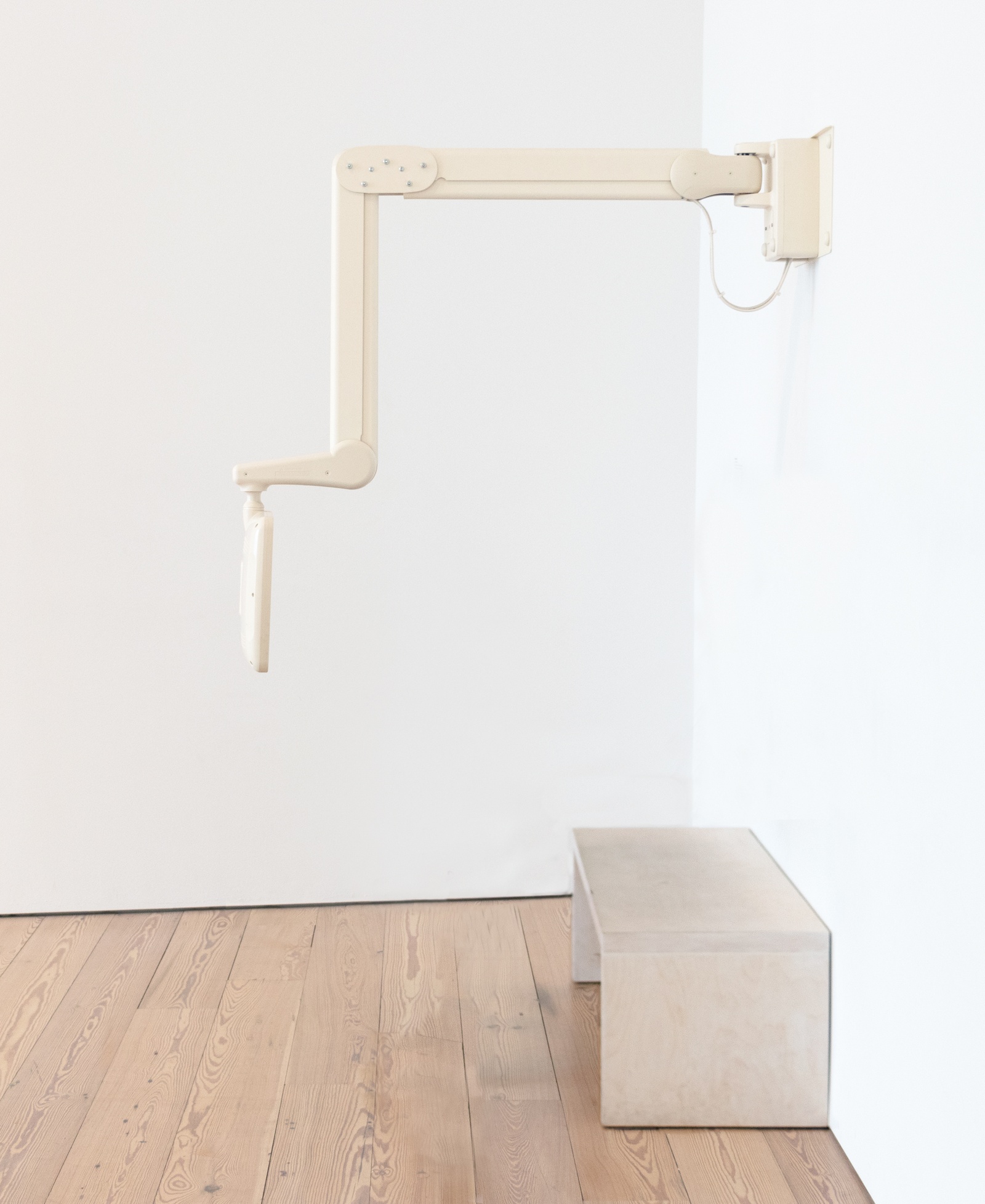 [In the corner of a white walls with a light wood floor, a beige mechanical arm mounted perpendicular to the wall extends outward holding a monitor. Below it, a simple wooden bench sits against the wall. The monitor is facing the wall and the bench.]