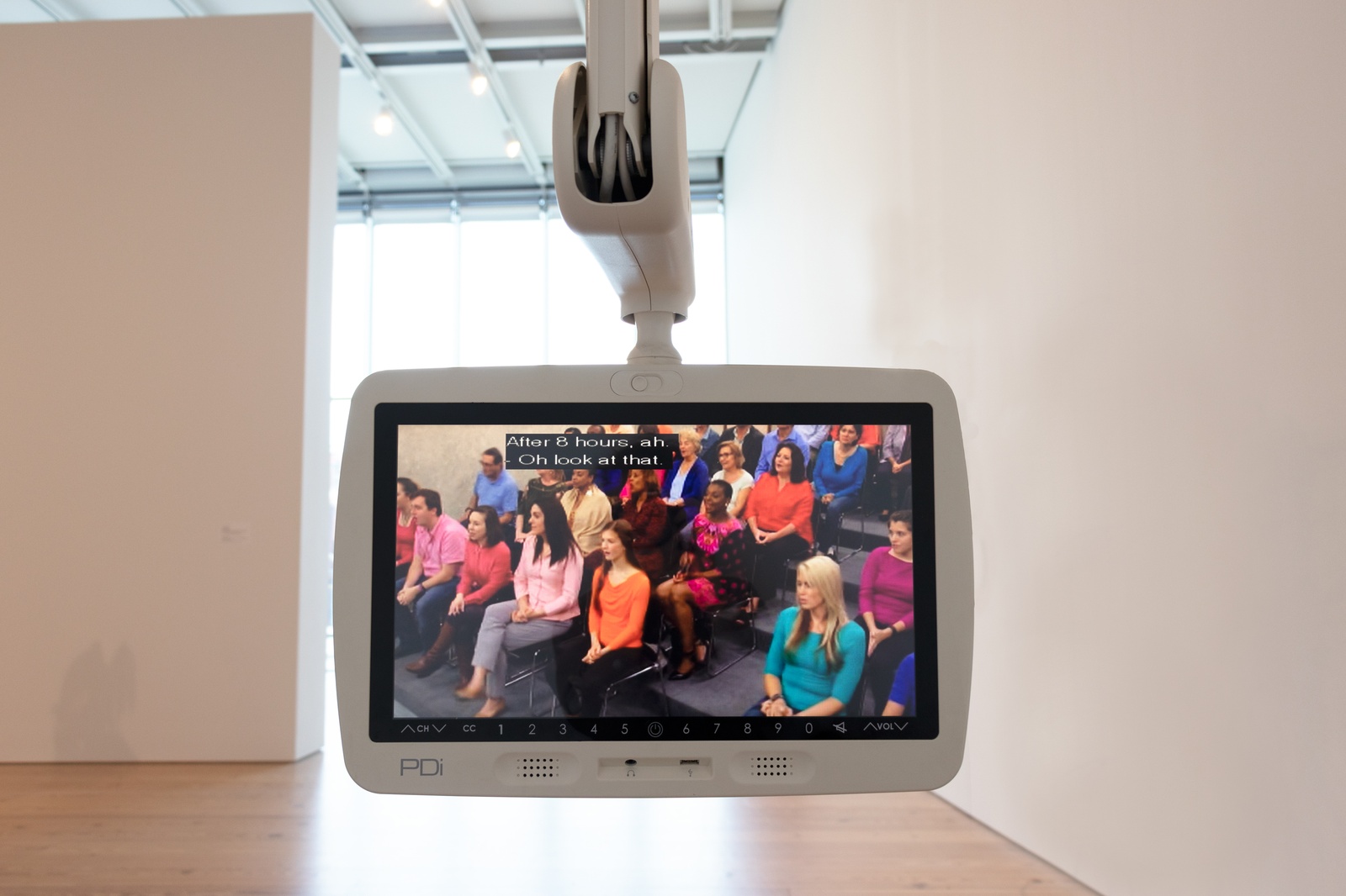 [A close-up of a hospital television monitor suspended from the ceiling by an articulated arm in a museum space. The background features white walls, light hardwood floors, and strong light streaming through windows. The screen displays a cable TV broadcast of an audience seated in rows. They are casually dressed and reacting with surprise or curiosity to something off-camera. Overlaid on the video is text that reads, &quot;After 8 hours, ah. Oh look at that.&quot;]