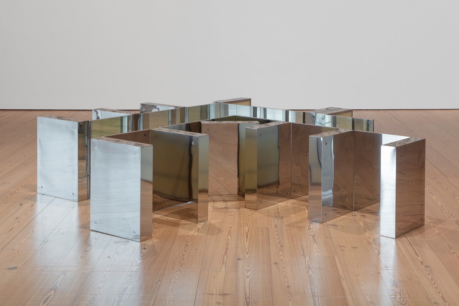 [In an exhibition space, a geometric sculpture consists of several mirrored medicine cabinets, each with a matte translucent surface on the inside. The cabinets are arranged in a zigzag pattern on the wooden floor. The mirrors reflect each other, the texture of the floor and the surrounding light, creating subtle changes in the appearance of the space. The reflective surfaces distort the environment, making it feel dynamic and fluid.]