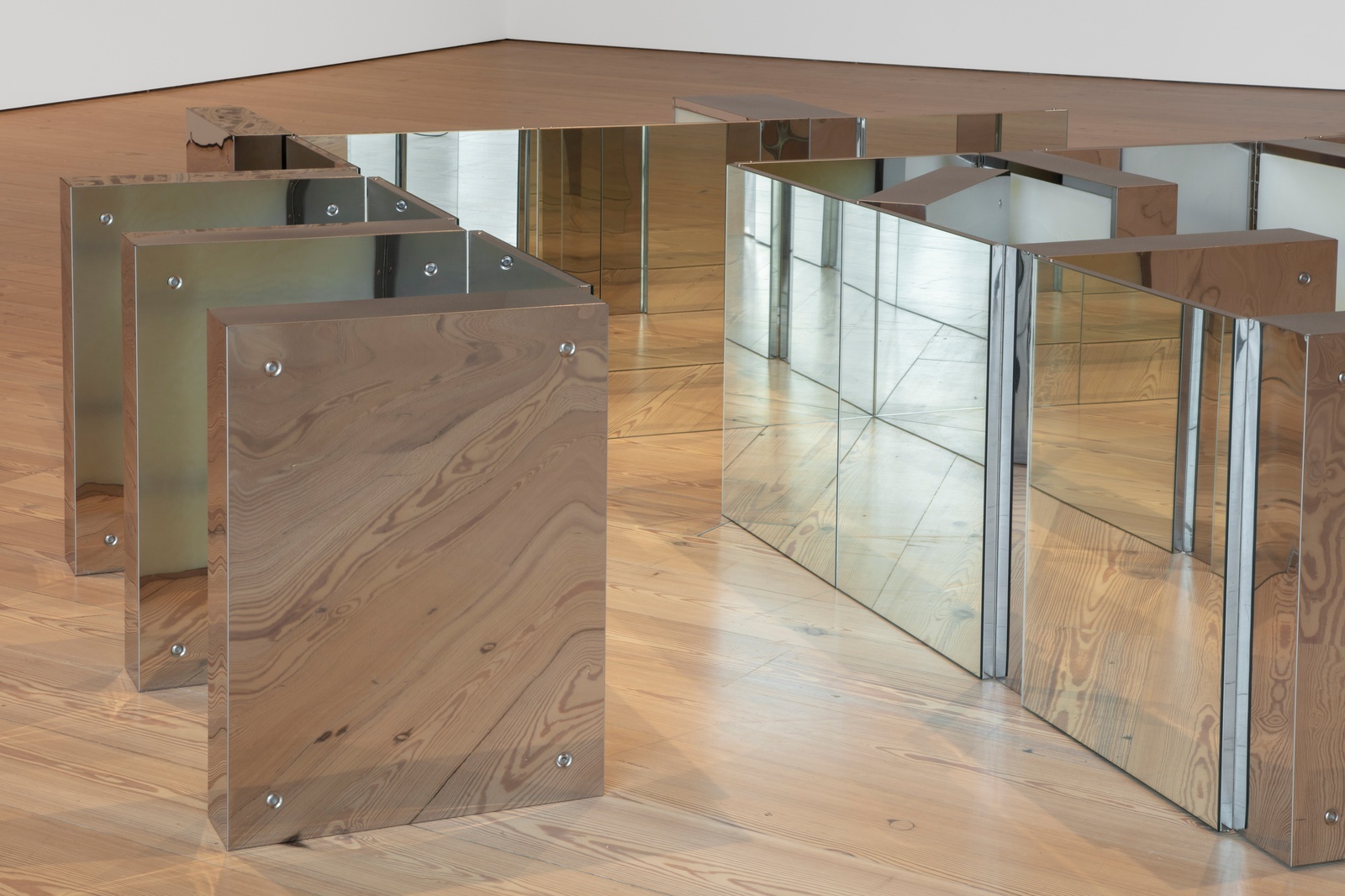 [Mirrored medicine cabinets are arranged in a zigzag pattern on a wooden floor. Some cabinets face outwards, reflecting the floor and surrounding space, while others are partially open, revealing a translucent, gel-like substance inside. The mirrored surfaces distort the reflections of the room, creating a sense of depth and fragmentation.]