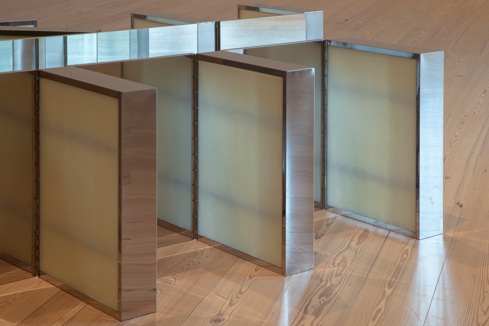 [The close-up captures a section of a larger geometric sculpture on a wooden floor, focusing on three medicine cabinets arranged in a grid-like formation. Their interiors are coated with a thick layer of Vaseline, creating a smooth, translucent surface that diffuses light and alters reflections.]