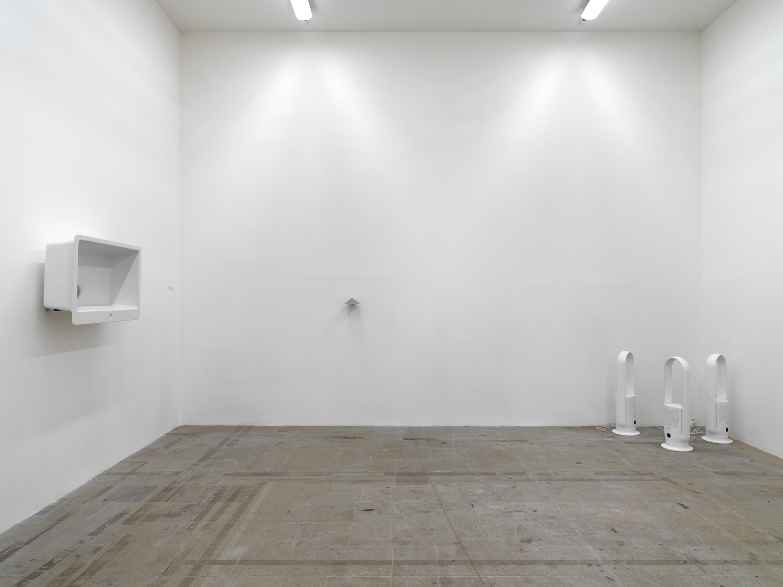 [On the left wall of the space a white ceramic sink hung vertically with its basin facing the center, approximating the overall shape and form of an old television. In the far right corner is a cluster of three standing air purifiers. In the center of the back wall a smaller object is hung at the wall.]