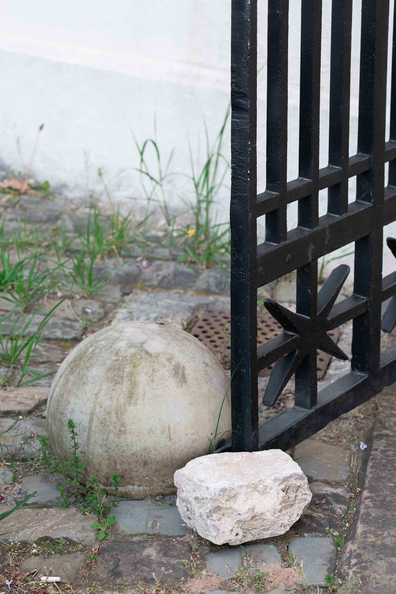 [A black wrought-iron gate entrance to an outdoor green space set between two white stone pillars. The gate features a decorative pattern with circular and pointed elements at the top. The gate is held open by a white natural stone. A narrow paved path leads through the gate into a green lawn. Behind the gate are bushes with purple lilac blossoms and dark green leaves. In the background, a white building with castle-like architectural details, including battlements and arched windows, is partially visible behind trees. The surrounding area features neatly trimmed grass and bushes. The scene appears to depict the entrance to a private estate, garden, or historical site.]