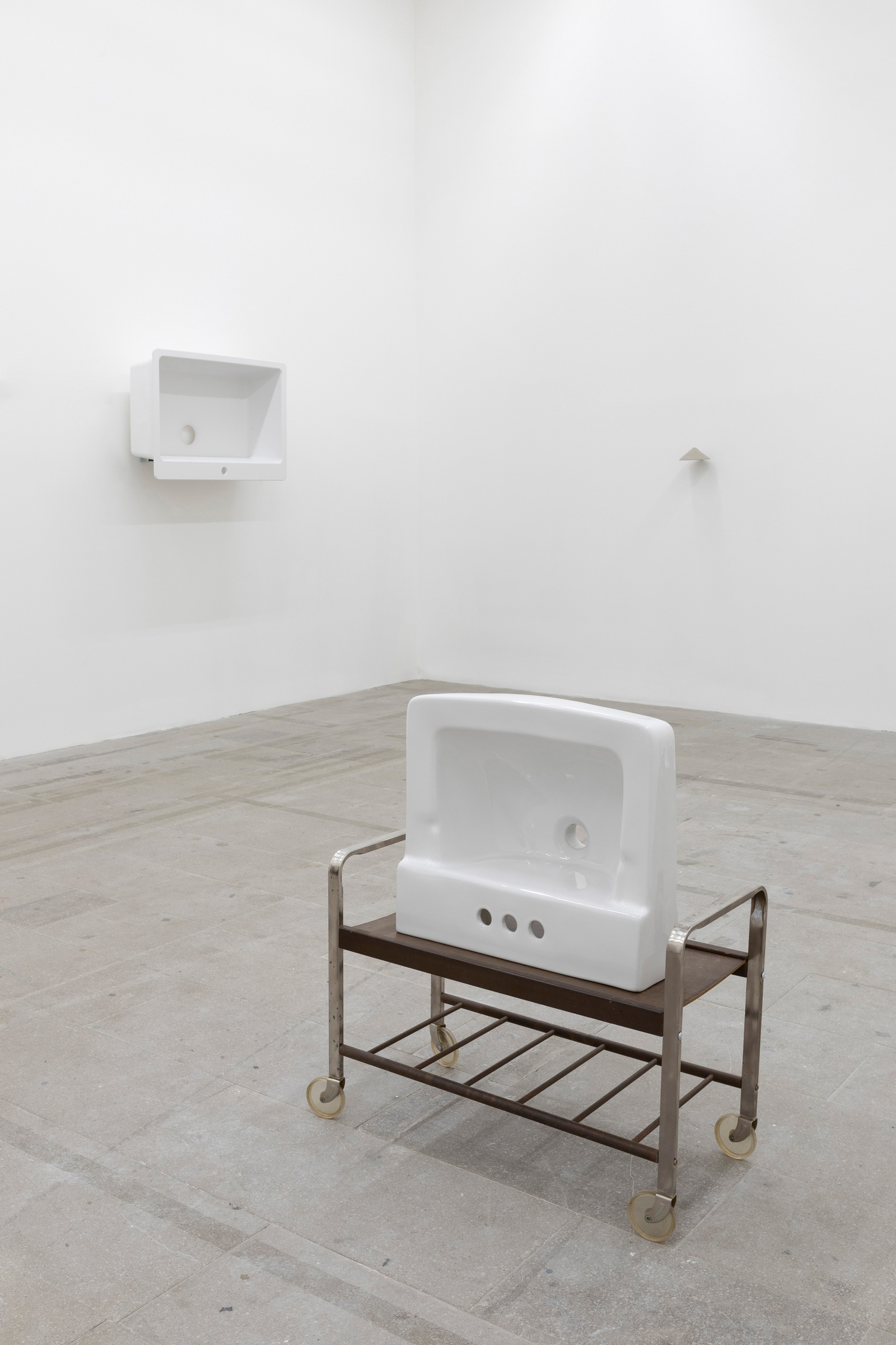 [In the foreground, is a white ceramic sink that sits upright atop a brown stand with wheels. The sink, with its basin facing forward, approximates the overall shape and form of an old television. In the far left corner is a white ceramic sink hung vertically on the wall with its basin facing forward, approximating the shape and form of an old television. On the right wall a smaller object is hung on the wall as well.]