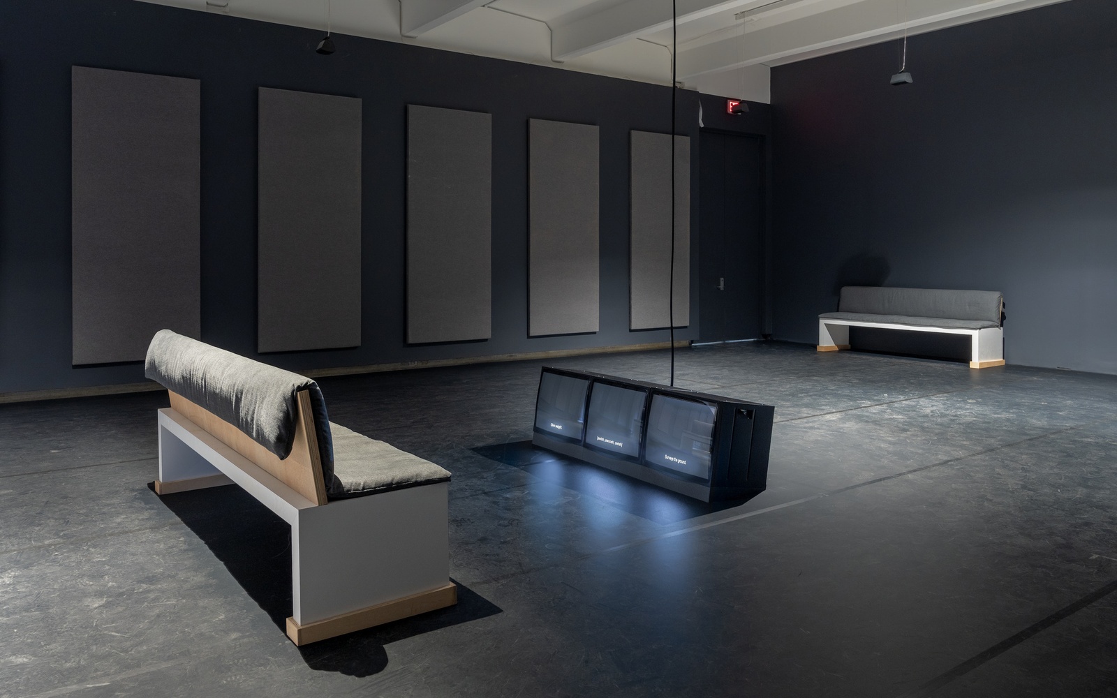 [An exhibition space is dimly lit by two spot lights. Two benches with wooden bases and cushioned tops are placed in the space, inviting visitors to sit and engage with the central installation. In the center of the room, a rectangular structure with three luminous television screens is placed on the floor, facing one of the benches. The screens emit a soft light that contrasts with the dark surroundings. Large rectangular acoustic panels line the walls.]