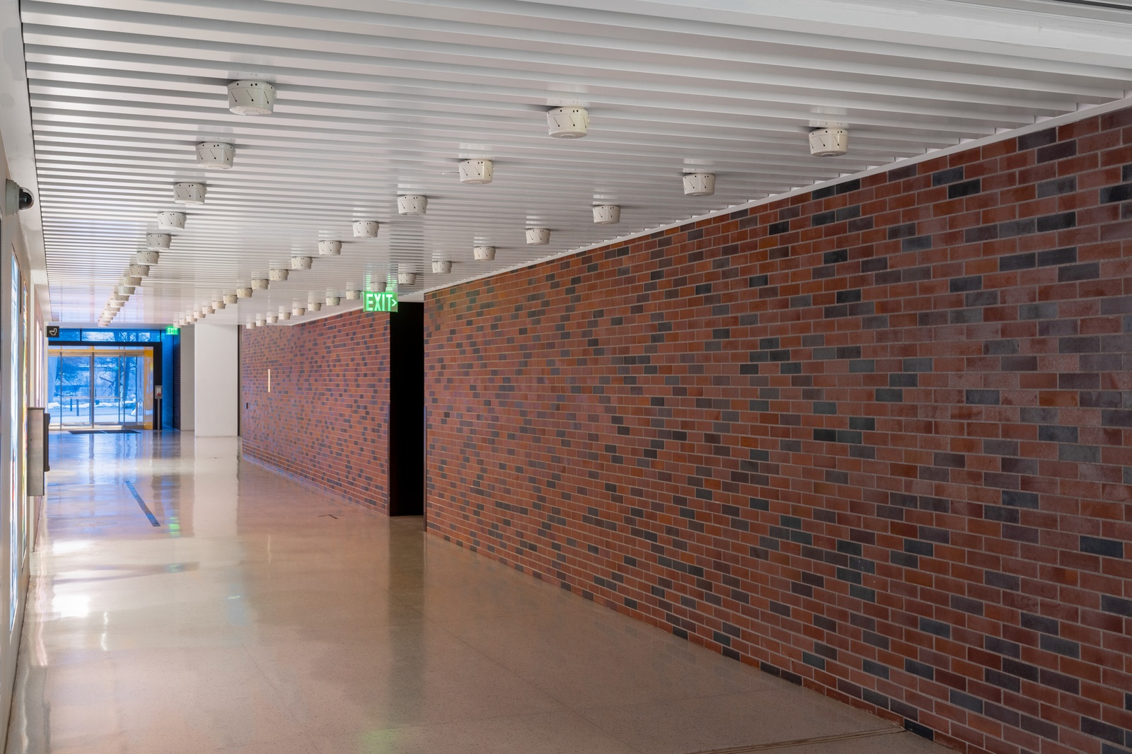 [The entrance hall features a white ceiling with jagged, horizontal edges, and three rows of sixteen smoke detectors are installed along it. To the right, there is a wall made of a mix of red and gray bricks, with a doorway in the center. Above the doorway, an LED exit sign glows with green light. At the end of the corridor, there is a windowed door.]
