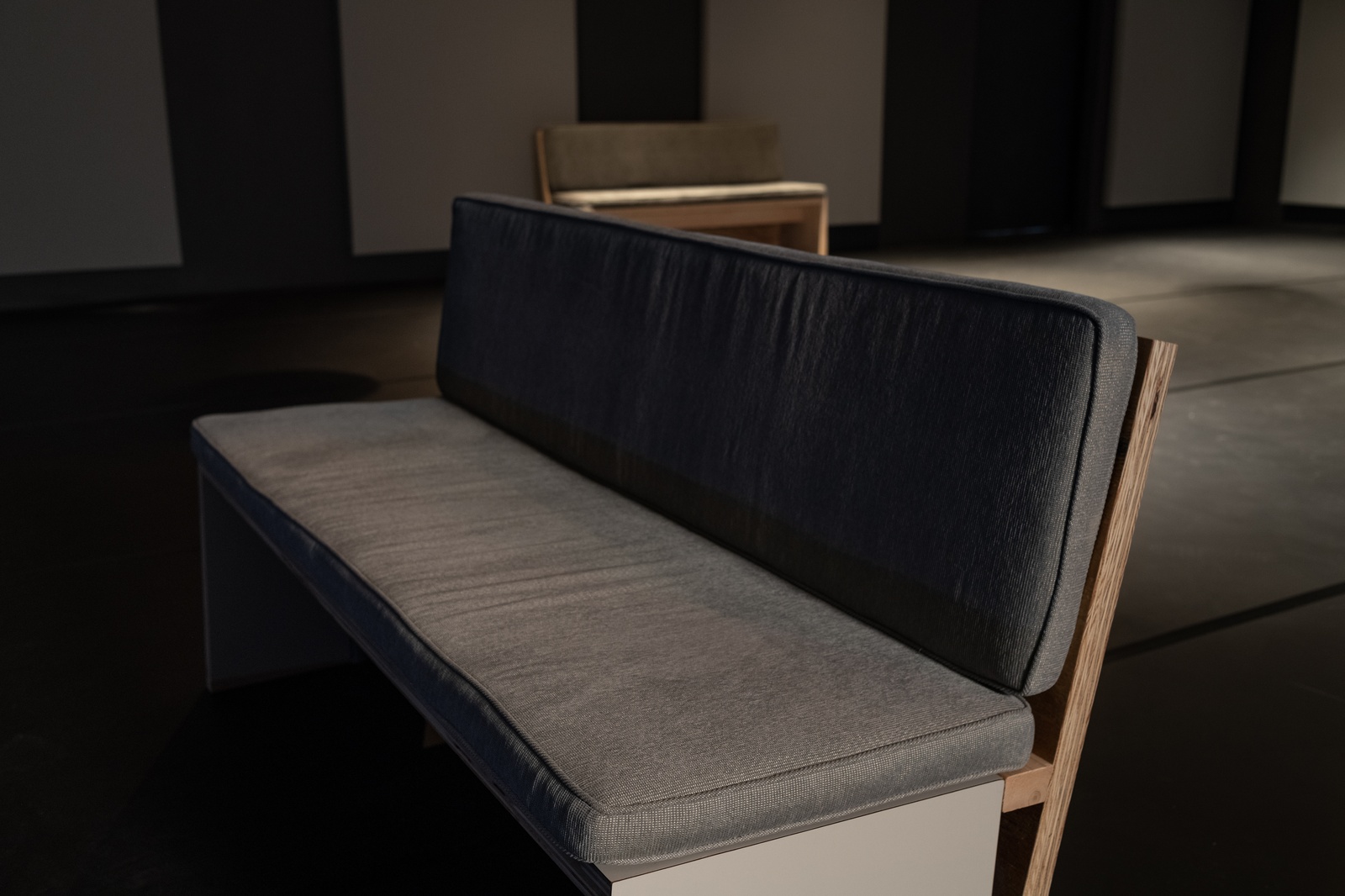[A close-up view of an exhibition bench is seen from the left side in a dark room. The bench has a wooden backrest and gray cushion. A second bench is visible in the background. The walls behind the bench are covered with acoustic panels.]